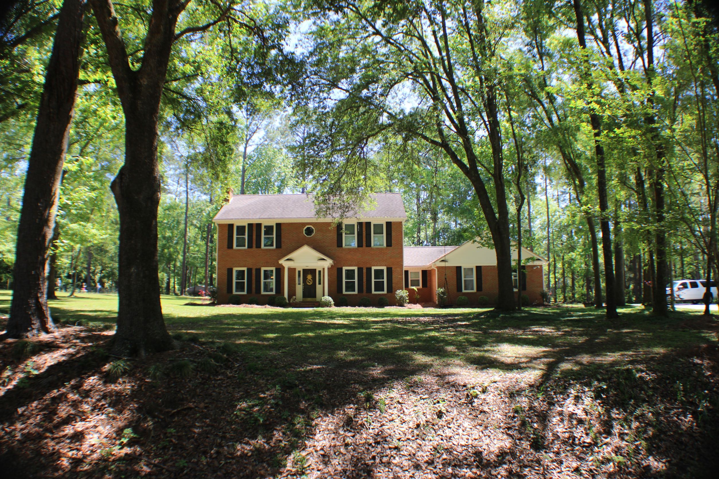 Pictured is a traditional red brick home that is squarish with black shutters. There is an attached garage and the photo is taken from a distance like the home has a large front yard. Home is found in Killearn Tallahassee Florida