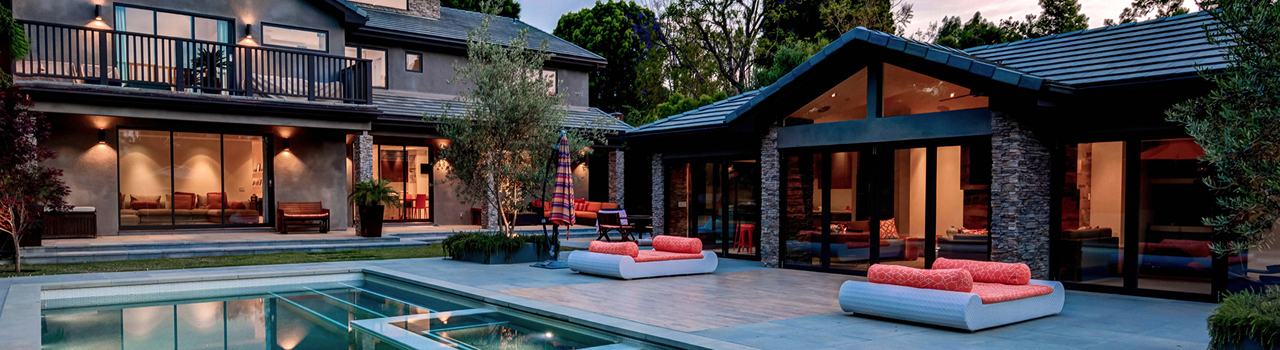 Decorative photo is of a patio and pool area that has been landscaped and lit with lights at night. The patio door is lit and the pool and area looks inviting.