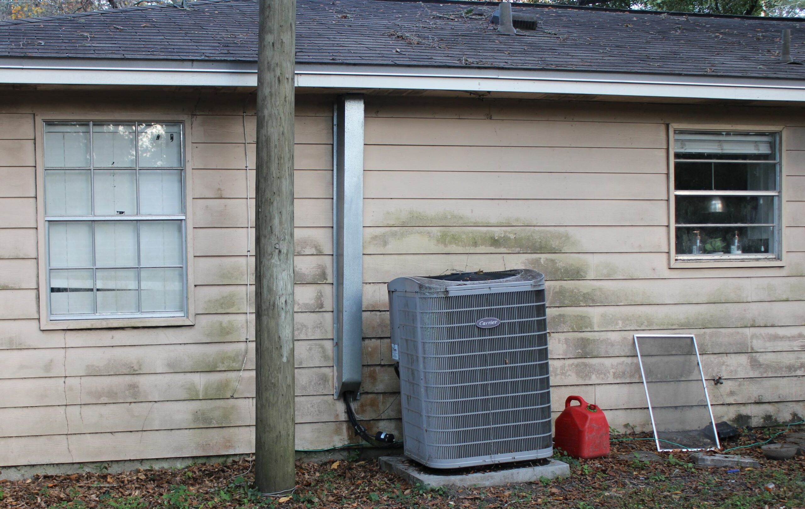 Photo shows back of a house including the kitchen window and the air conditioning unit. The siding is dirty with large dark patches. Window screen is leaning against the house rather than in the window.