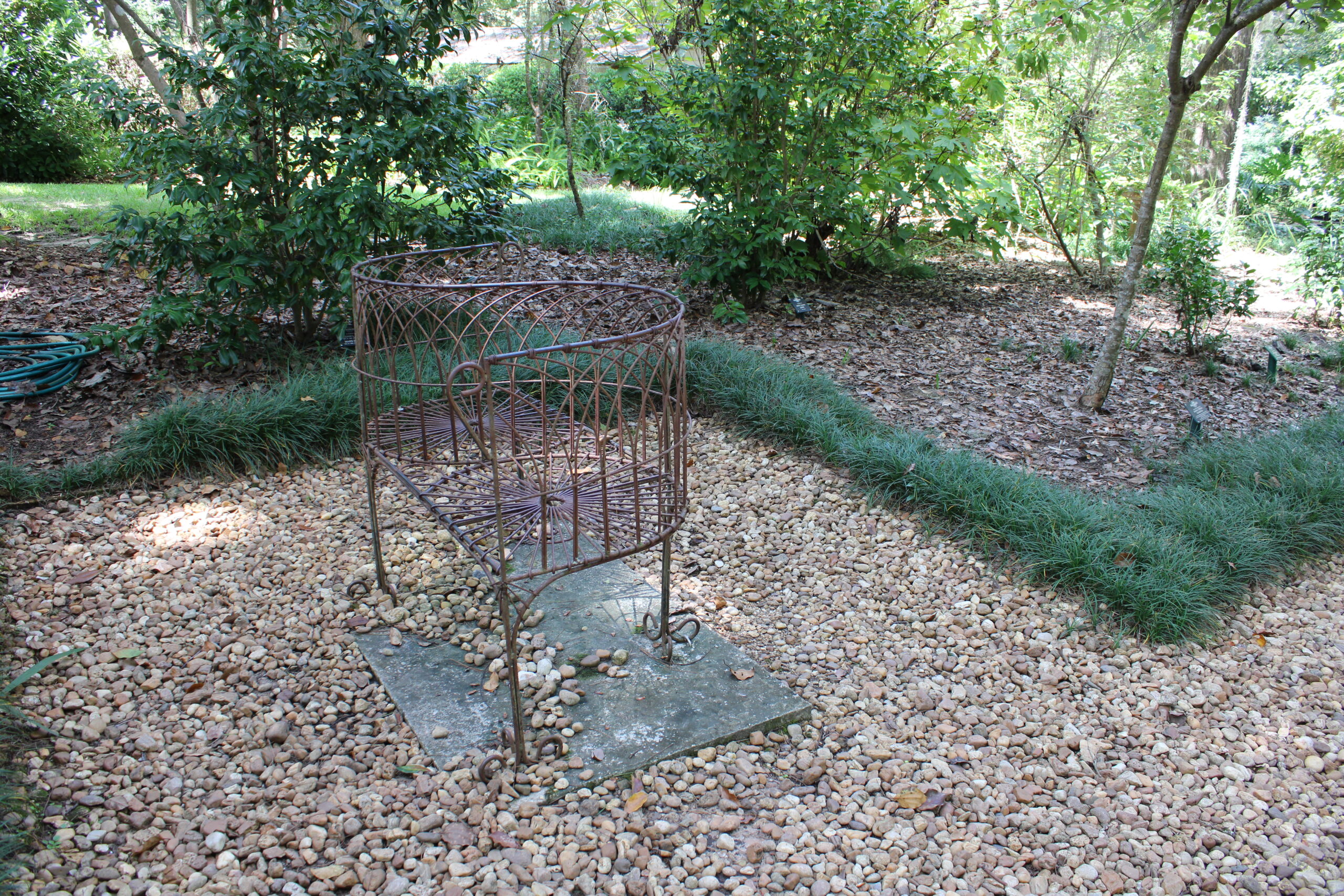 Decorative photo taken by Tallahassee Realtor. Shows a pebble walkway with a iron bench in the middle of a gravel patio. There are shrubs in the background and a edge made of green plants.